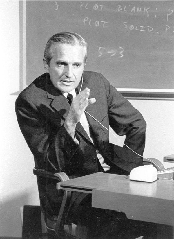 Douglas Engelbart at SRI in 1968, wearing a headset, gesturing, with the original wooden mouse on the desk beside him